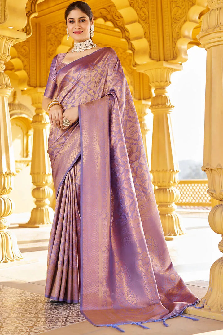 Woman in a purple saree with gold patterns standing in an ornate architectural setting.