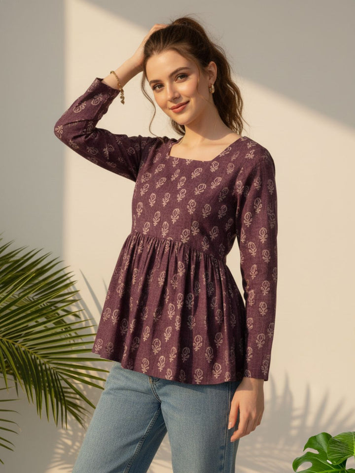 Woman wearing a purple patterned top and blue jeans standing against a light background with plants.