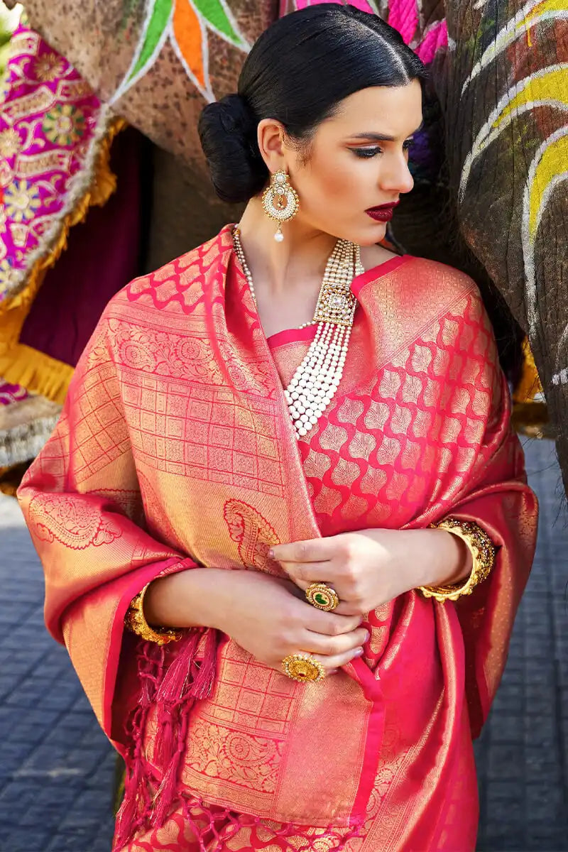 Woman in traditional pink and orange saree with jewelry, standing against a colorful background.