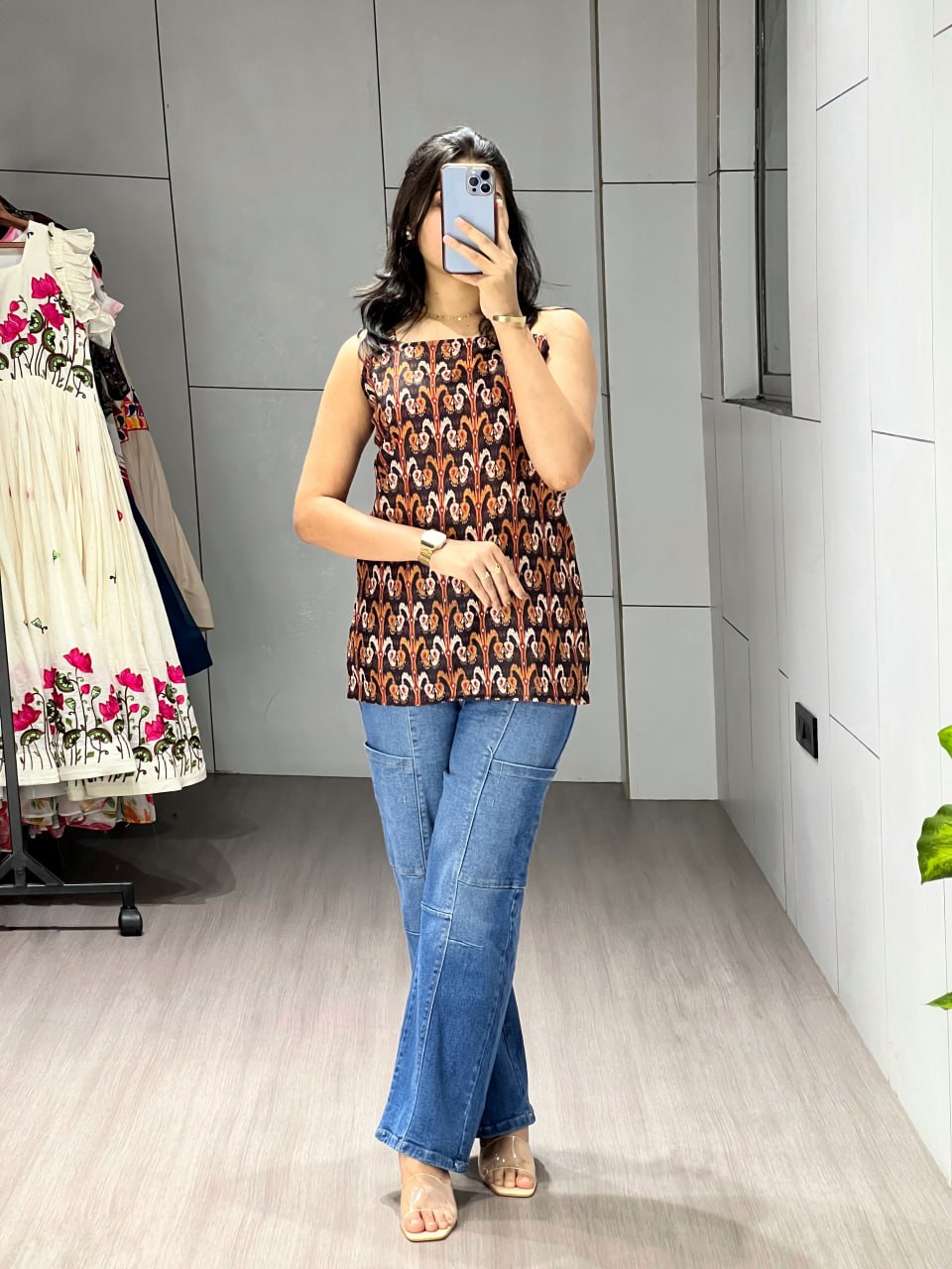 Person wearing a patterned top and blue jeans taking a mirror selfie in a room.