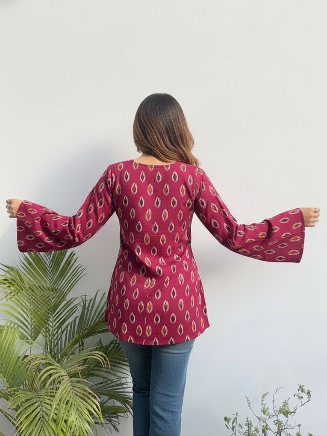 Person wearing a red patterned blouse with blue jeans, standing against a white wall with green plants.