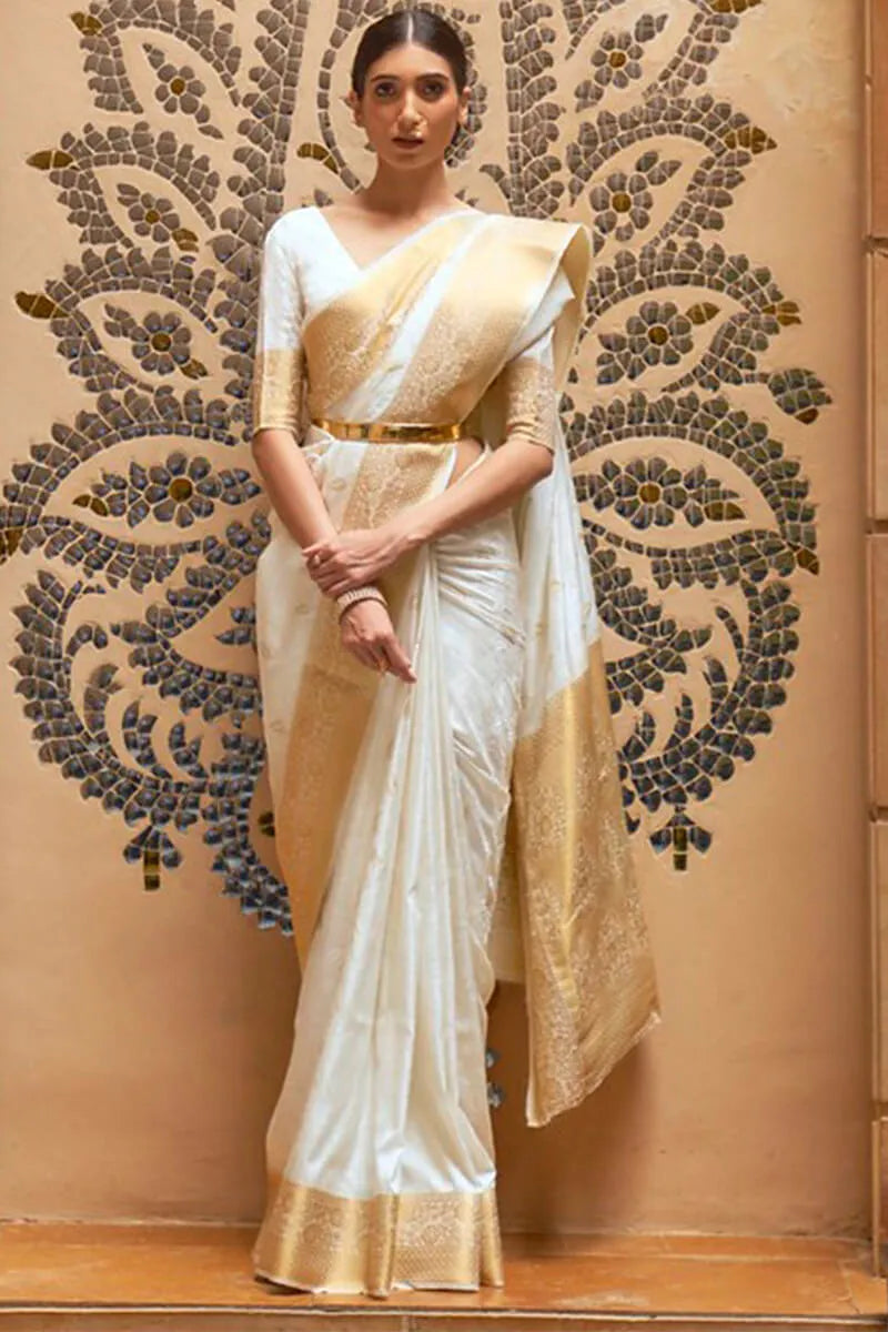 Woman wearing a white and gold saree against an ornate wall.