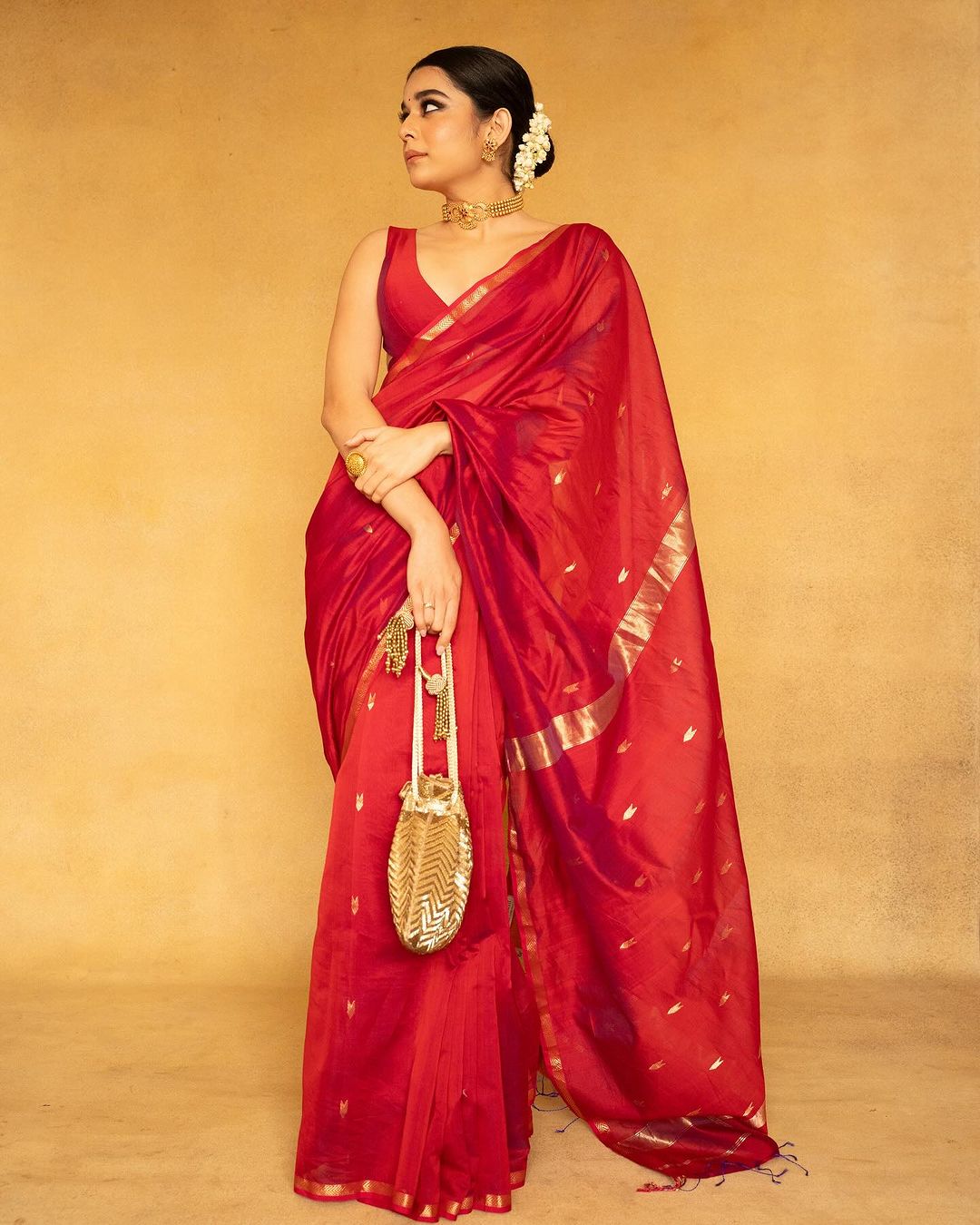 Woman in a red saree with gold accents holding a gold bag against
