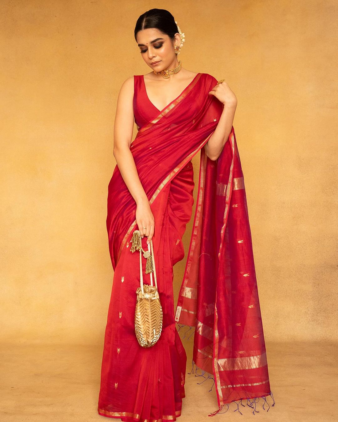 Woman in a red saree with gold accents holding a gold bag against