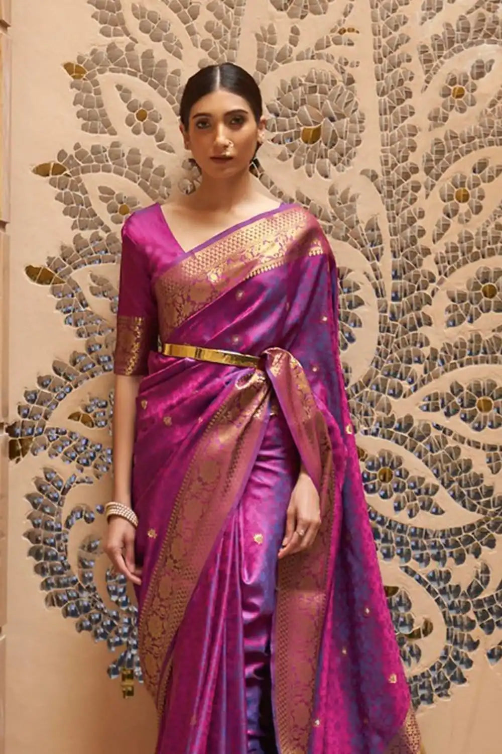 Woman wearing a pink saree with gold border against an ornate wall.