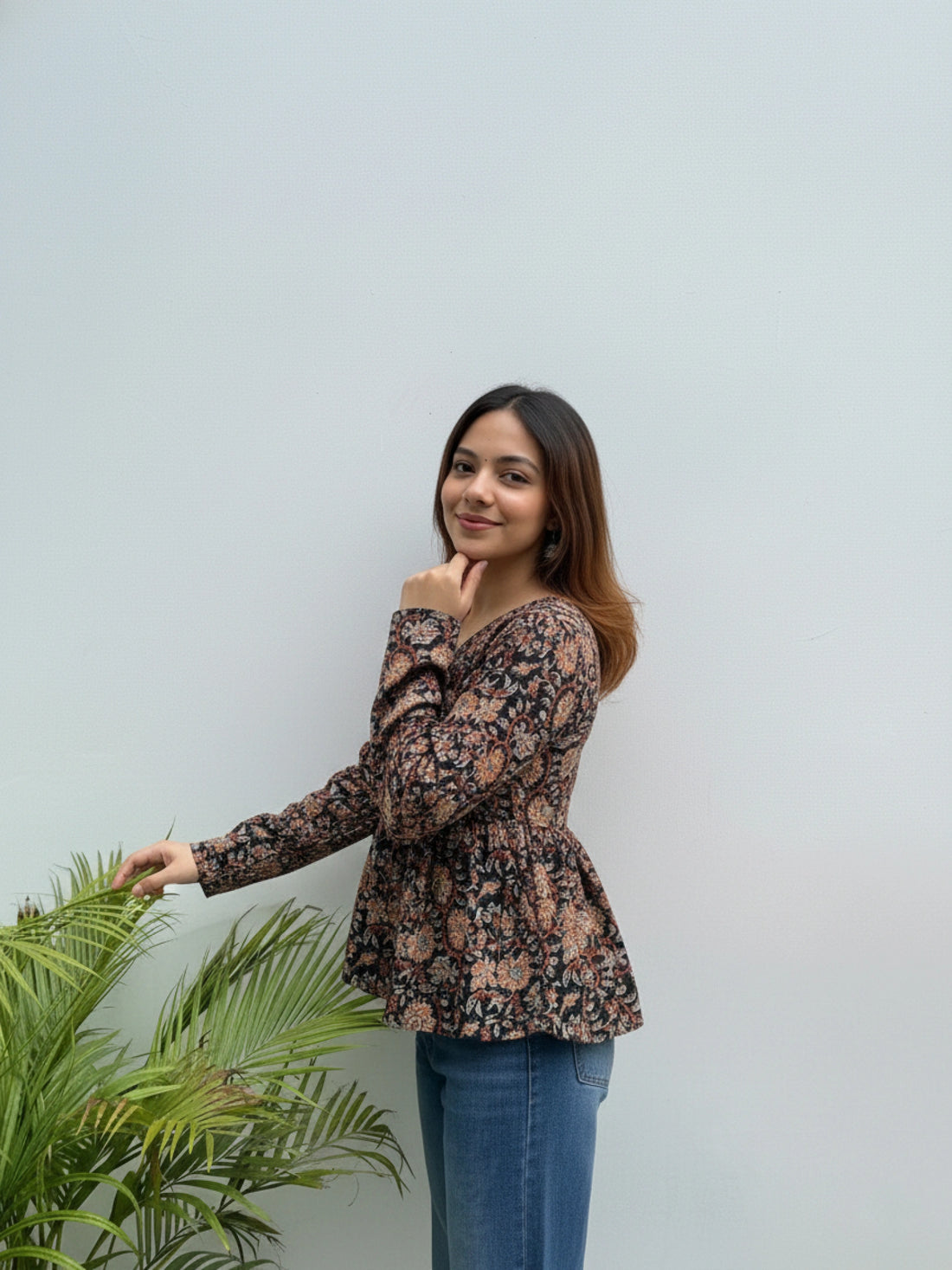 Woman wearing a floral blouse and blue jeans standing next to a plant against a light gray background