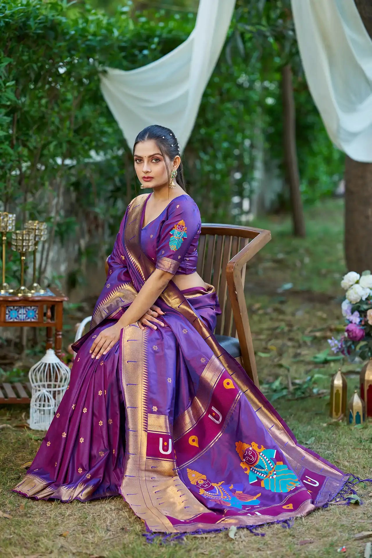 Woman in a purple saree with gold and blue patterns sitting outdoors.