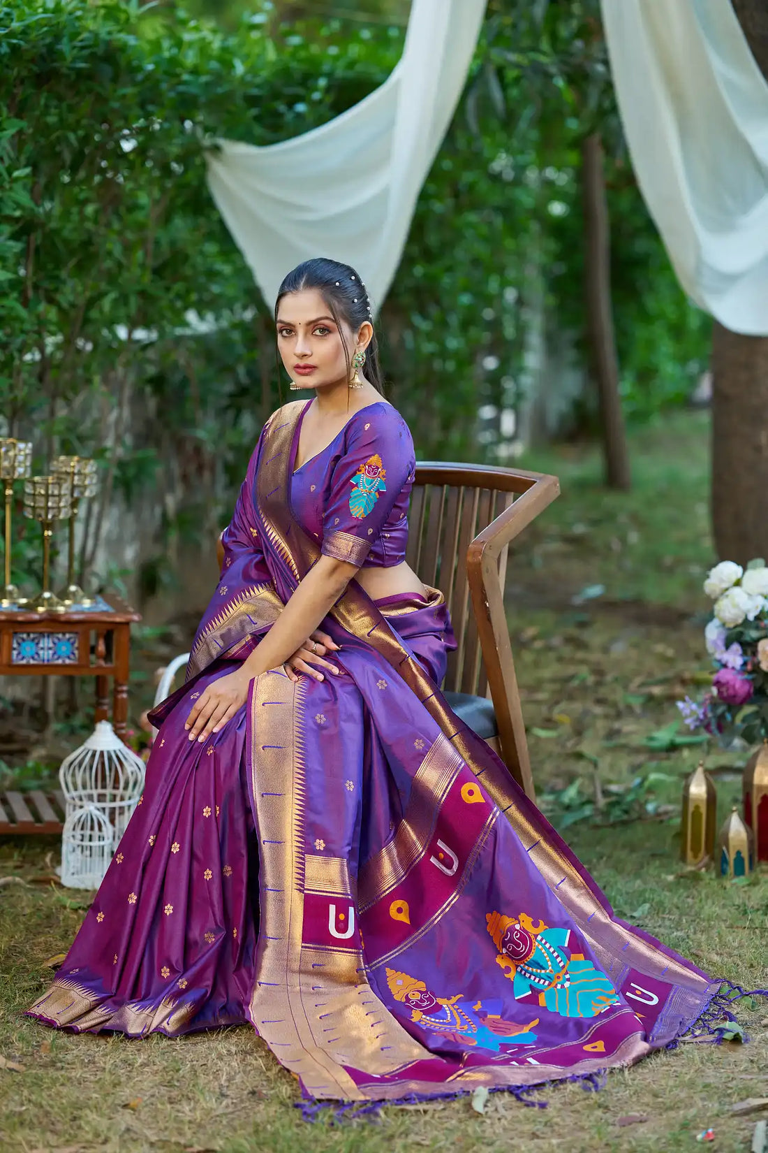 Woman in a purple saree with gold and blue patterns sitting outdoors.