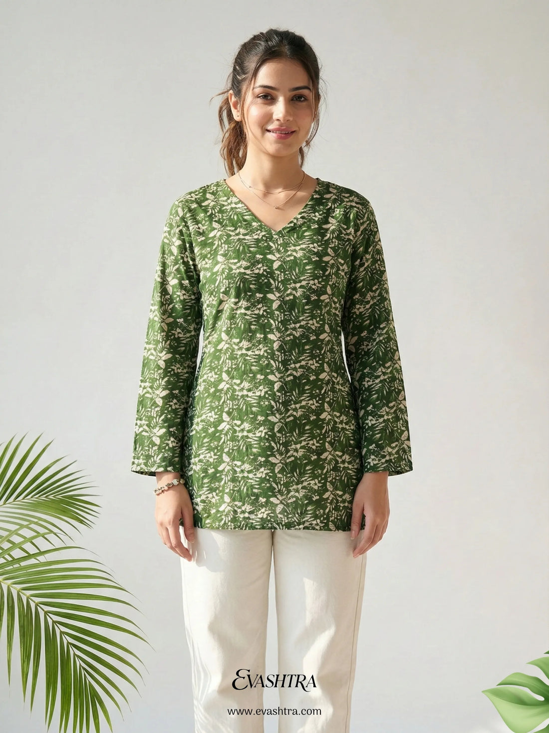 Woman wearing a green patterned top with white pants on a light gray background, featuring the brand 'Evashtra'.