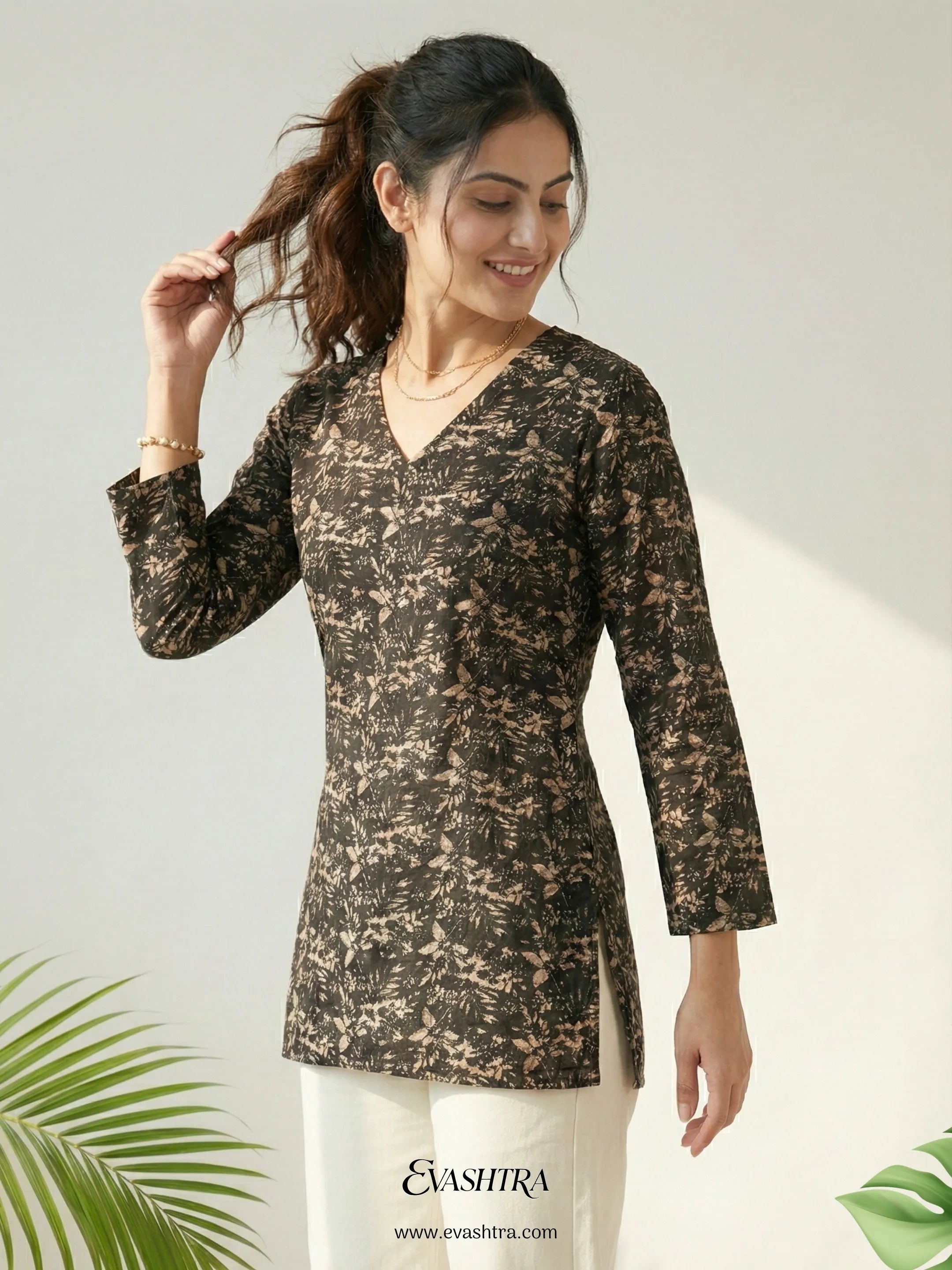 Woman wearing a black floral patterned top with white pants, standing against a light background with green leaves.