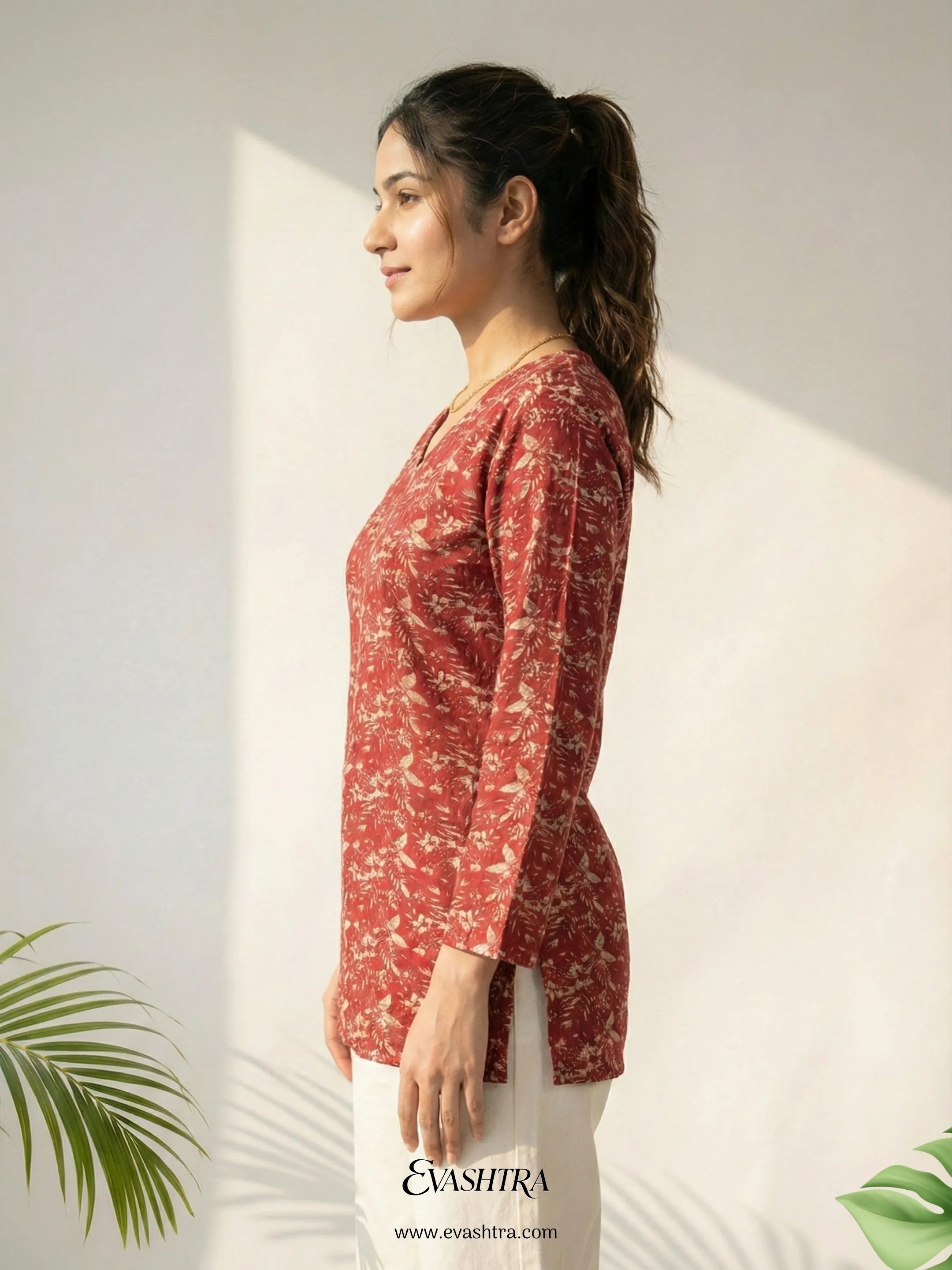 Woman wearing a red floral top against a white wall with a plant in the corner.