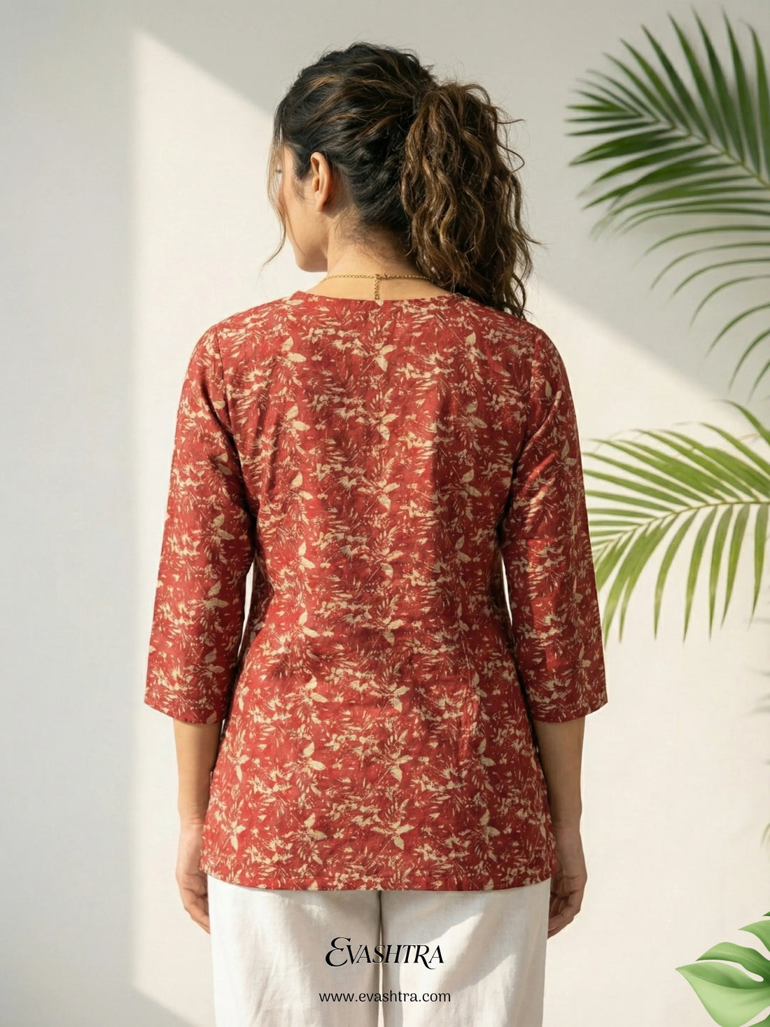 Woman wearing a red patterned top with white pants, standing against a light background with green leaves.