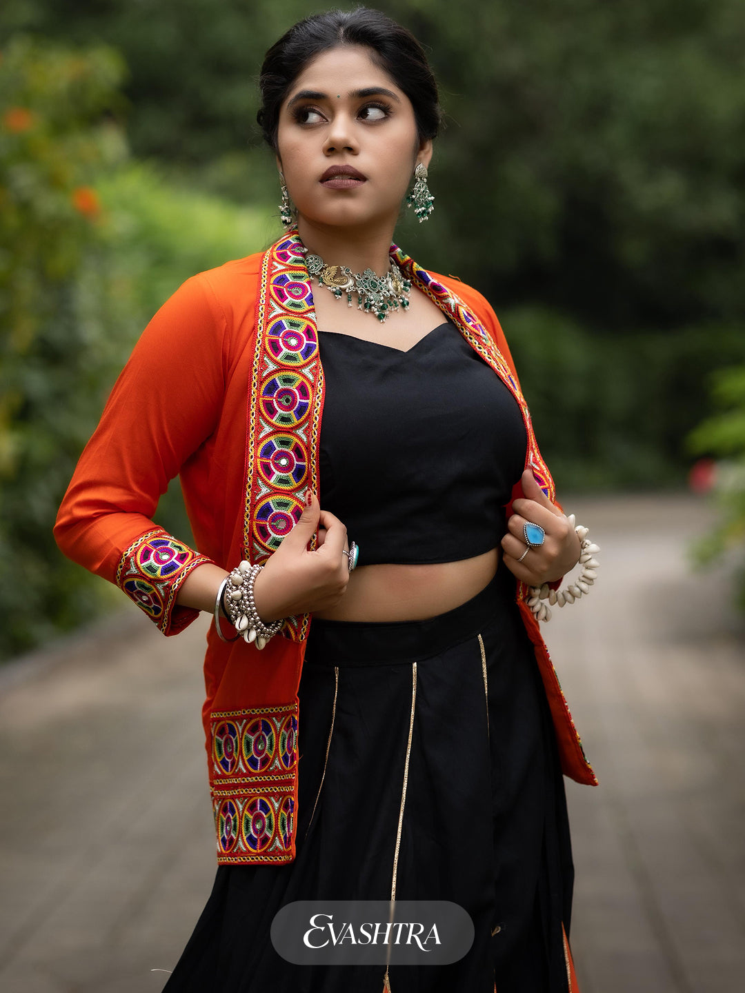 Woman wearing a traditional outfit with a colorful jacket and black saree, standing outdoors.
