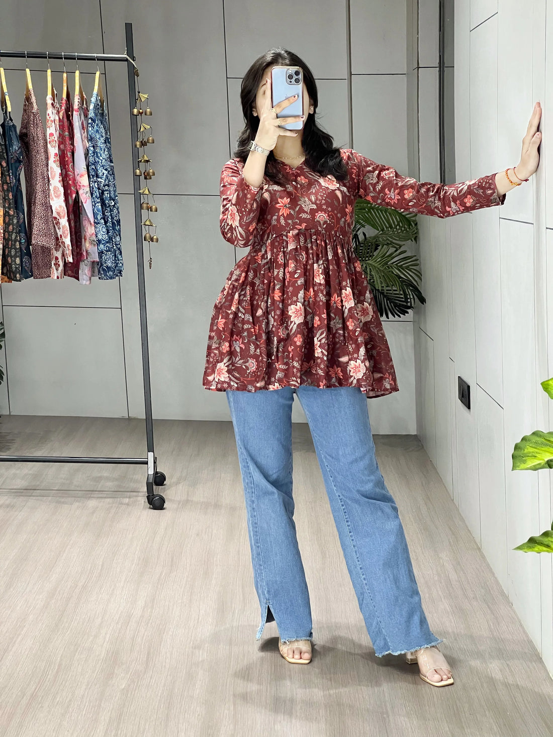 Person taking a mirror selfie wearing a floral top and blue jeans in a room with a clothing rack.