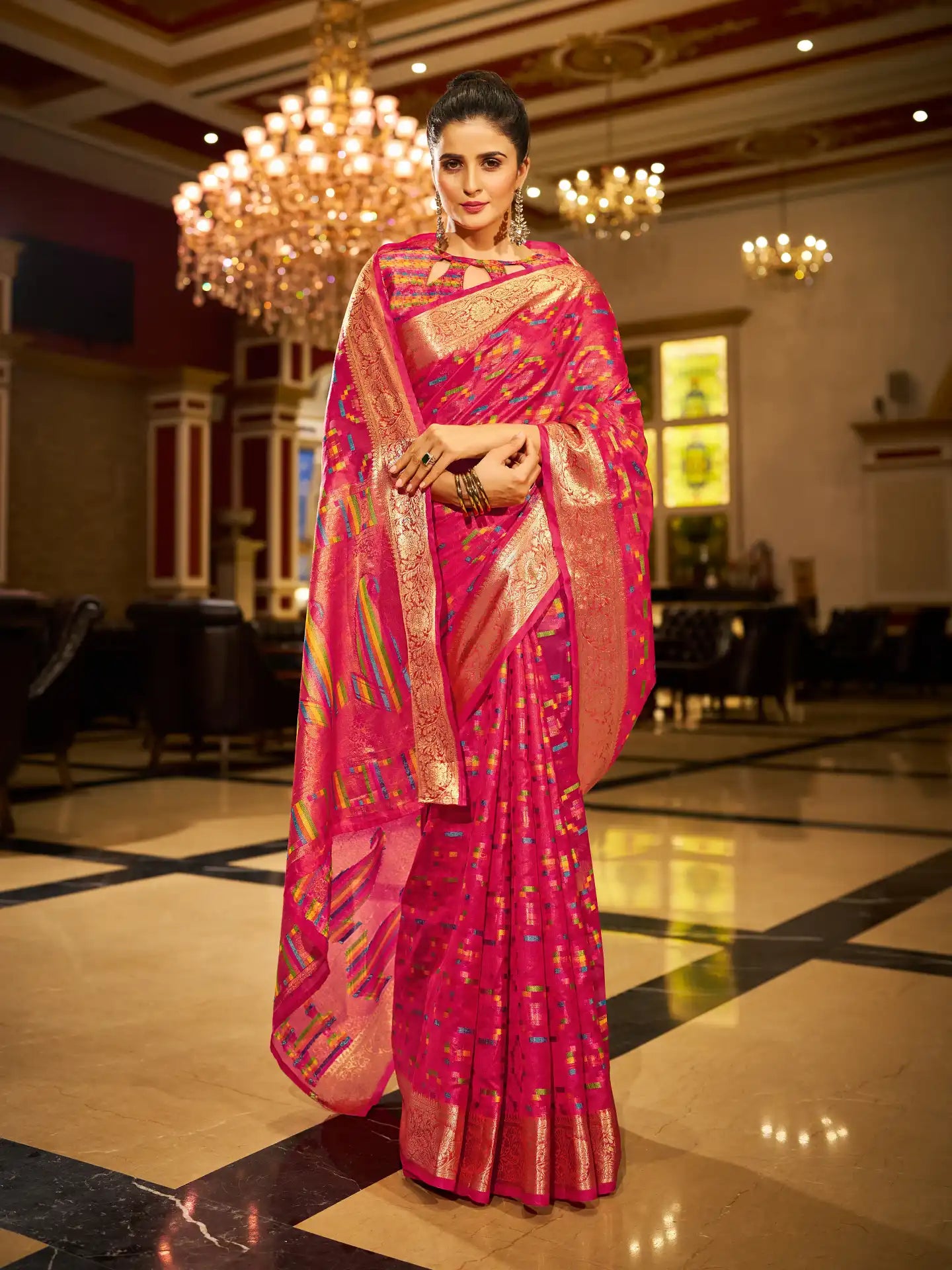 Woman in a pink saree with gold patterns standing in an elegant indoor setting.