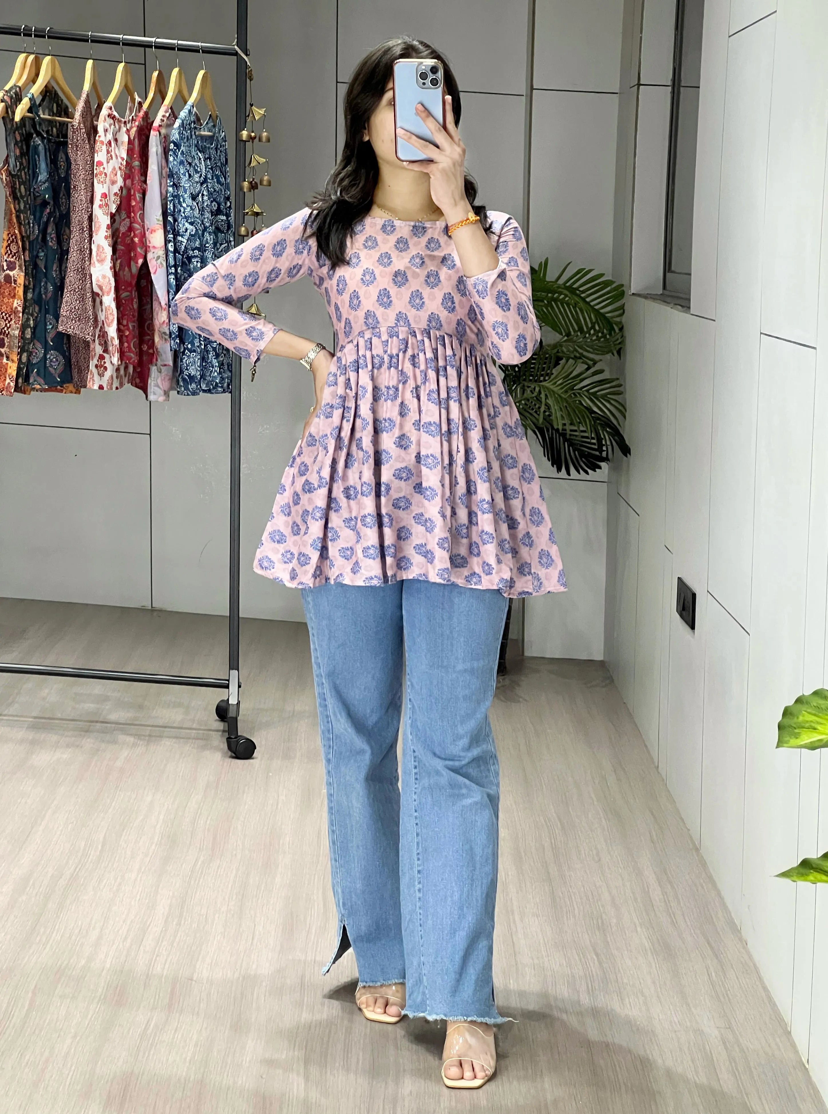 Woman taking a mirror selfie wearing a floral top and blue jeans in a room with a rack of clothes in the background.