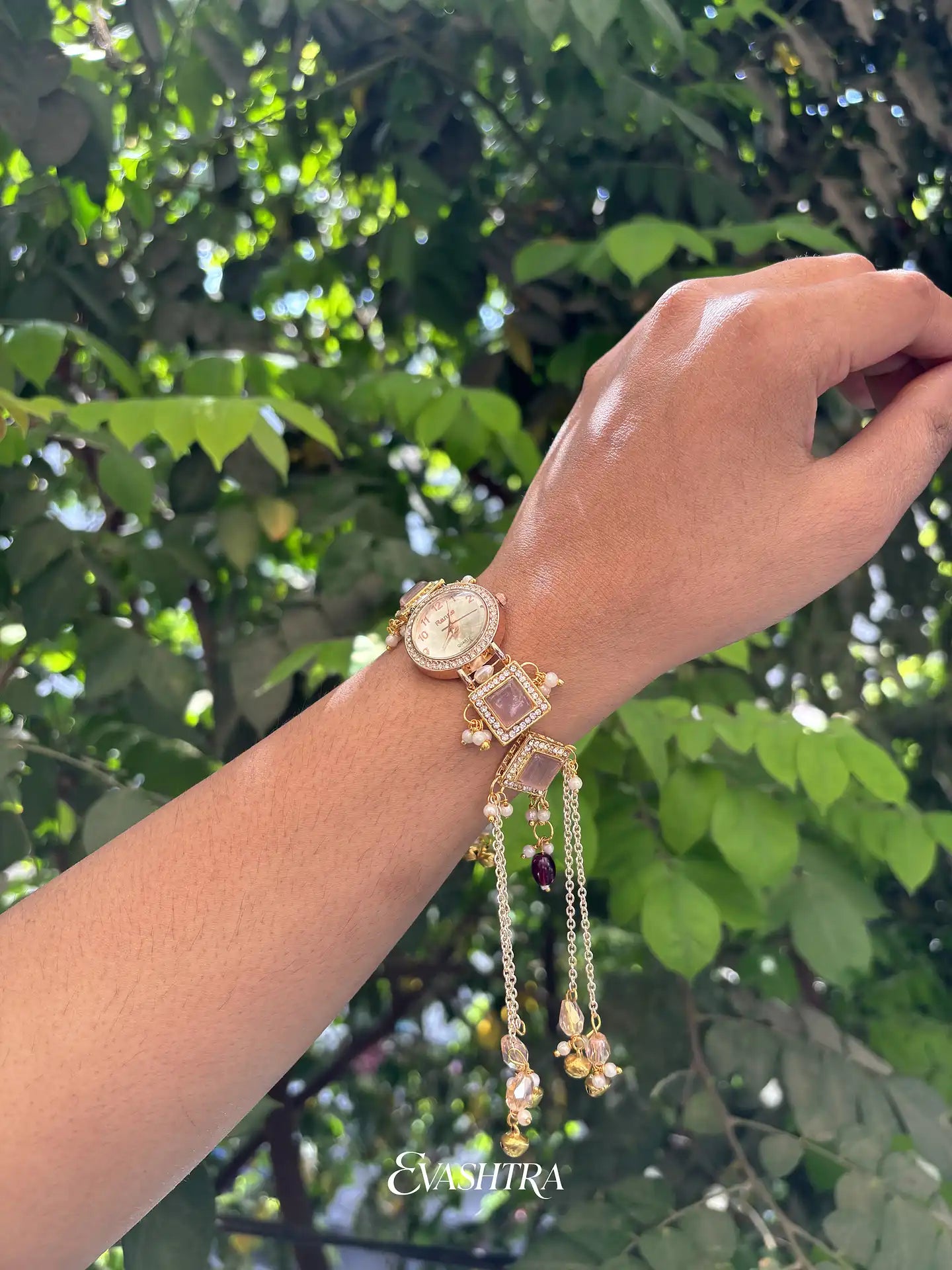 Hand wearing a gold watch and bracelet with a green leafy background