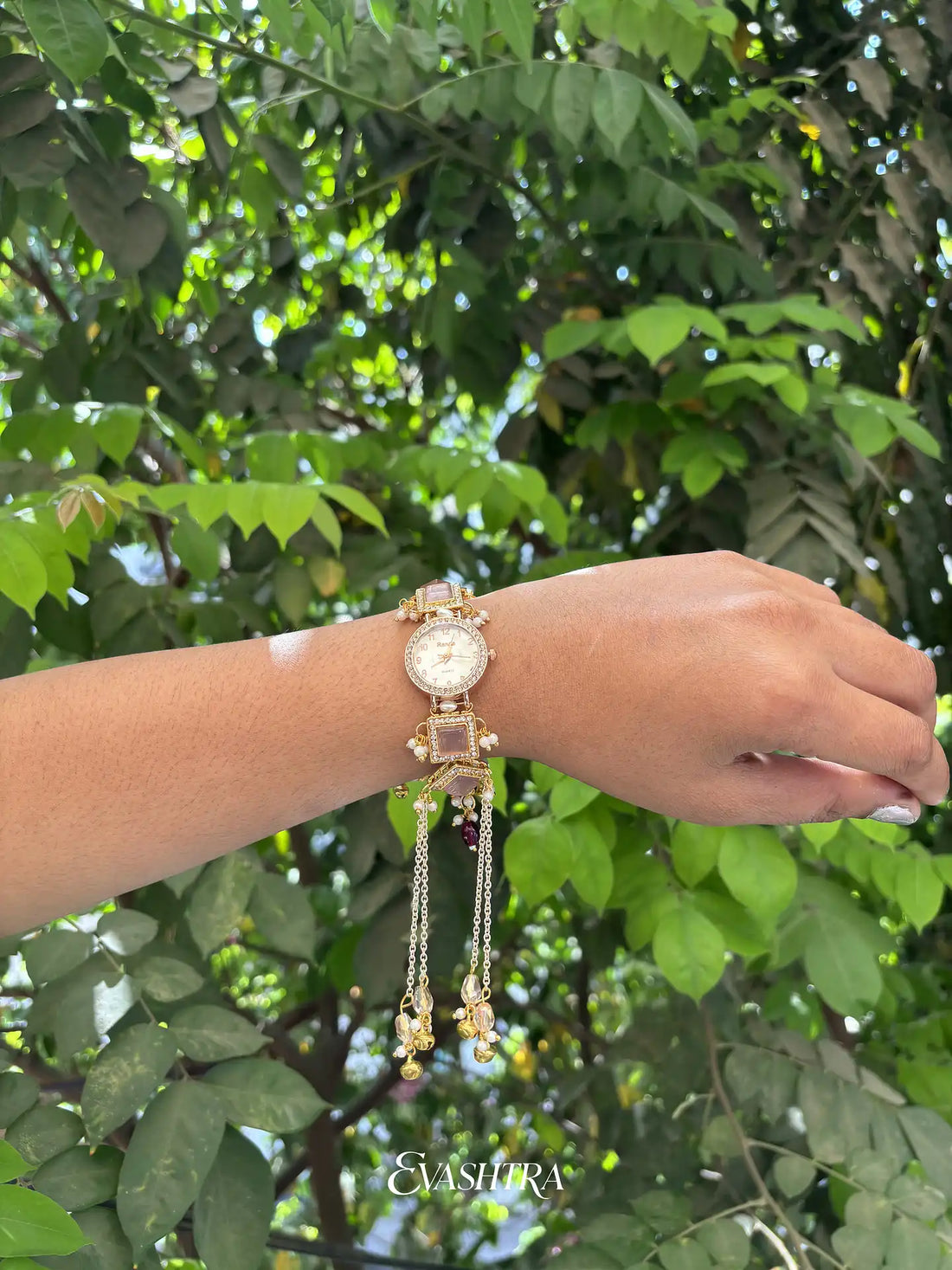 Hand wearing a gold bracelet with a watch face against a green leafy background