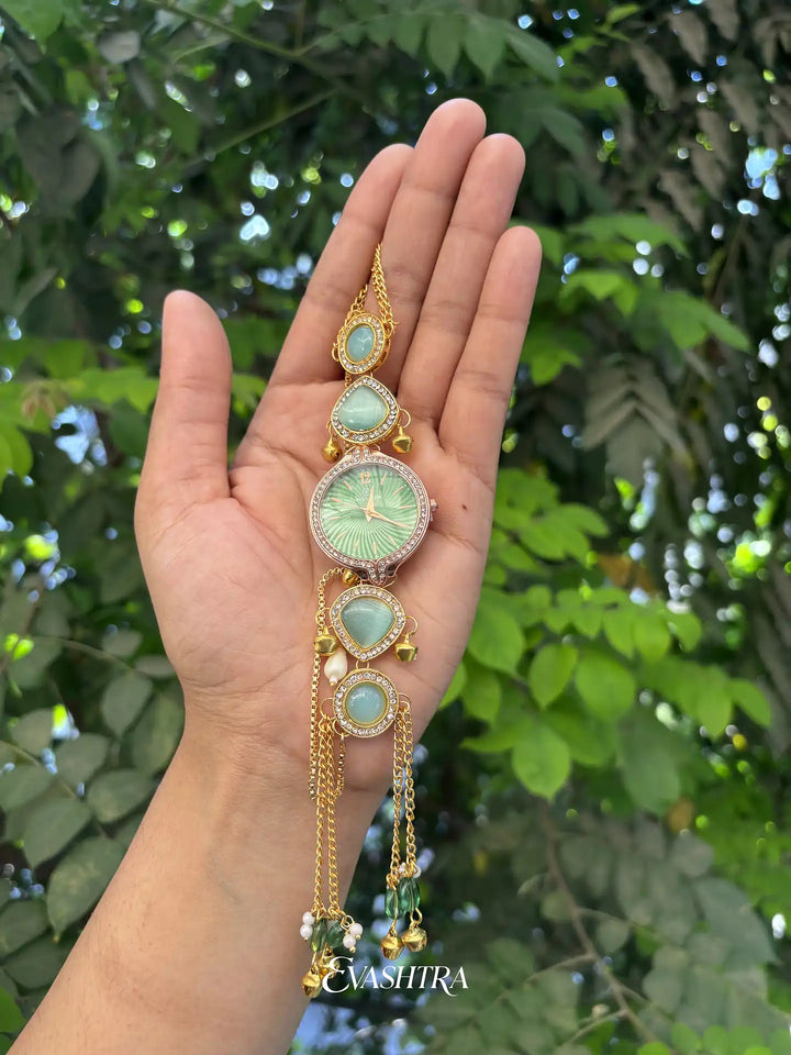 Hand holding a decorative watch with green gemstones against a leafy background