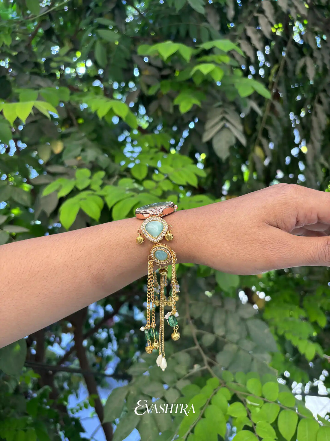 Bracelet with green gemstone and gold accents on a wrist against a green leafy background, featuring the brand 'Evashira'.