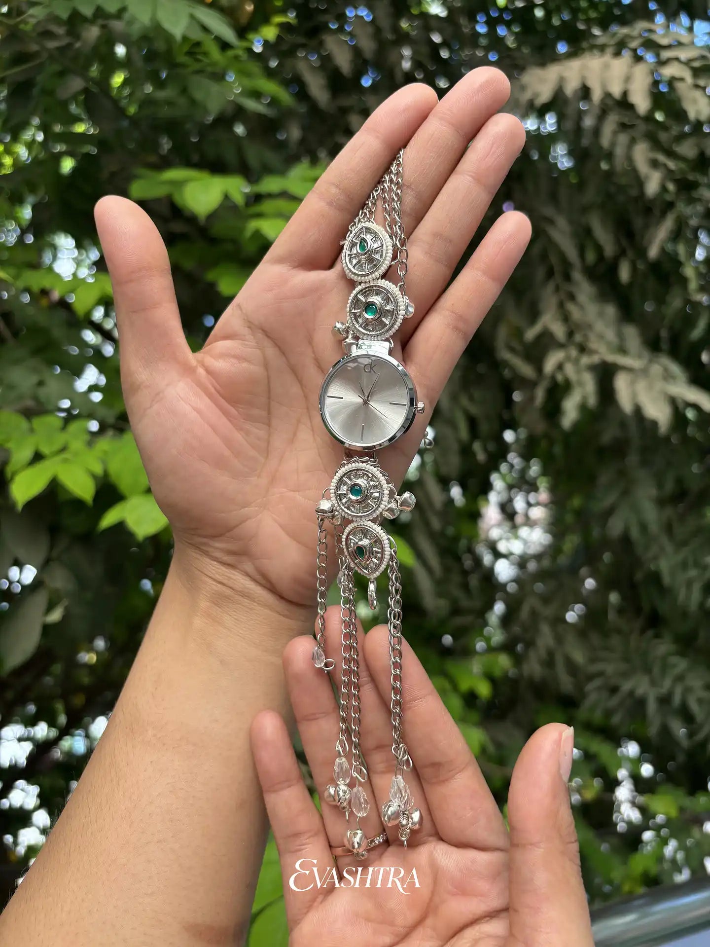 Silver bracelet with a watch face held in a hand against a green leafy background