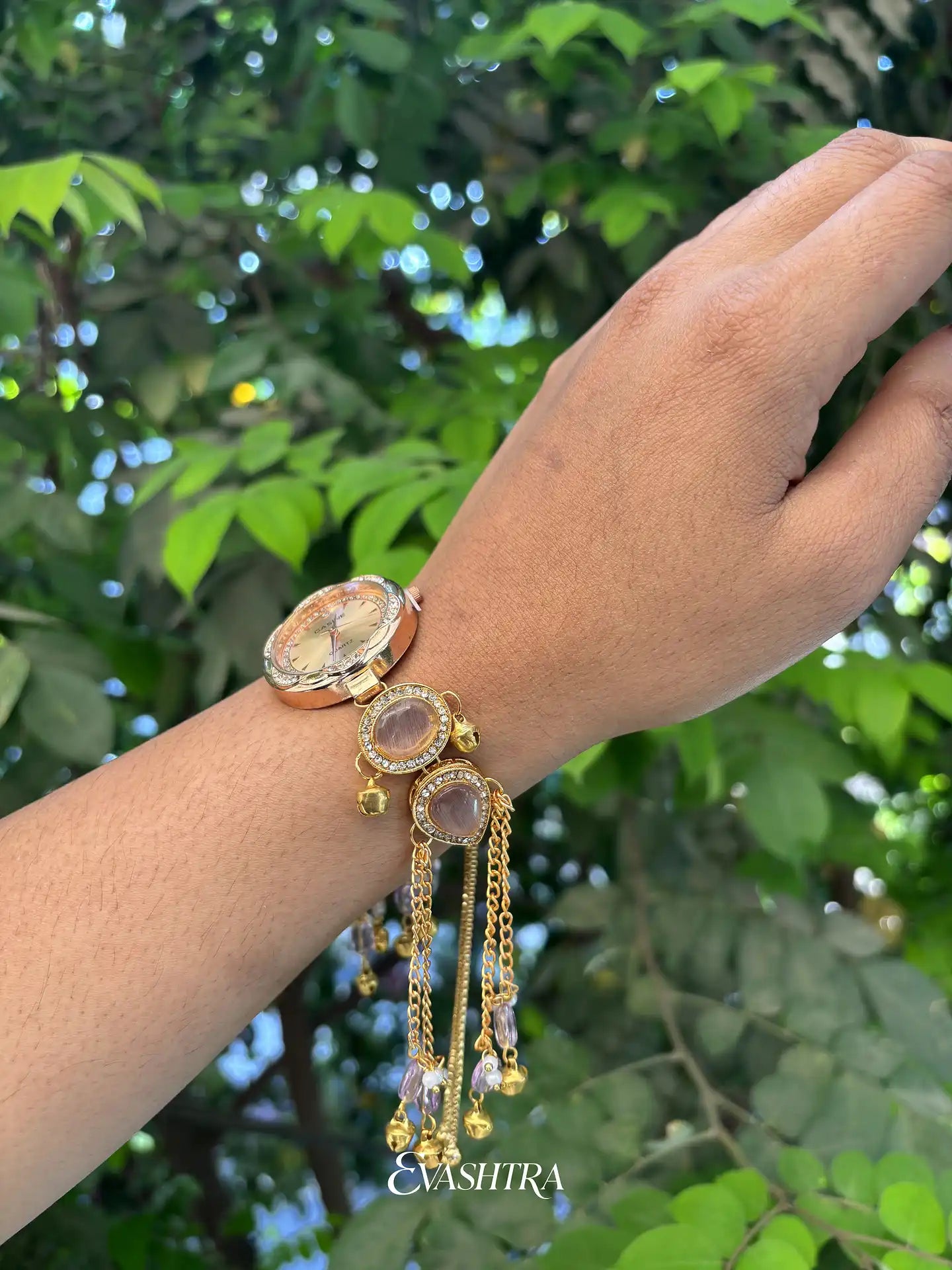 Hand wearing a gold bracelet with gemstones against a green leafy background