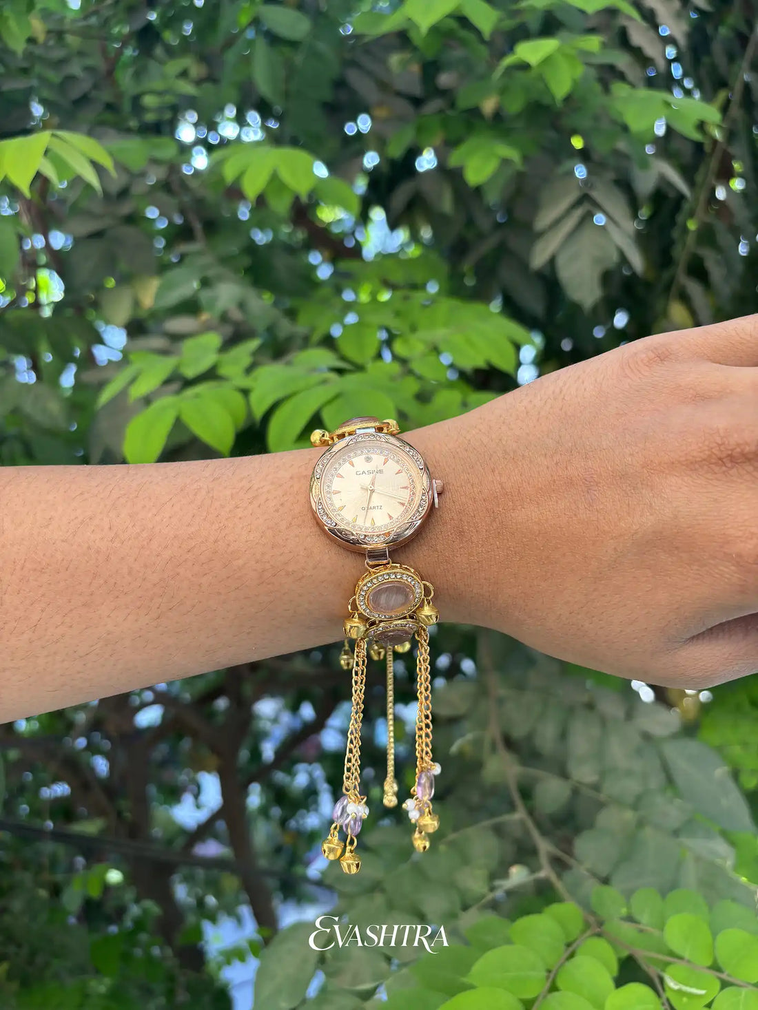Gold wristwatch with dangling elements on a wrist against a green leafy background