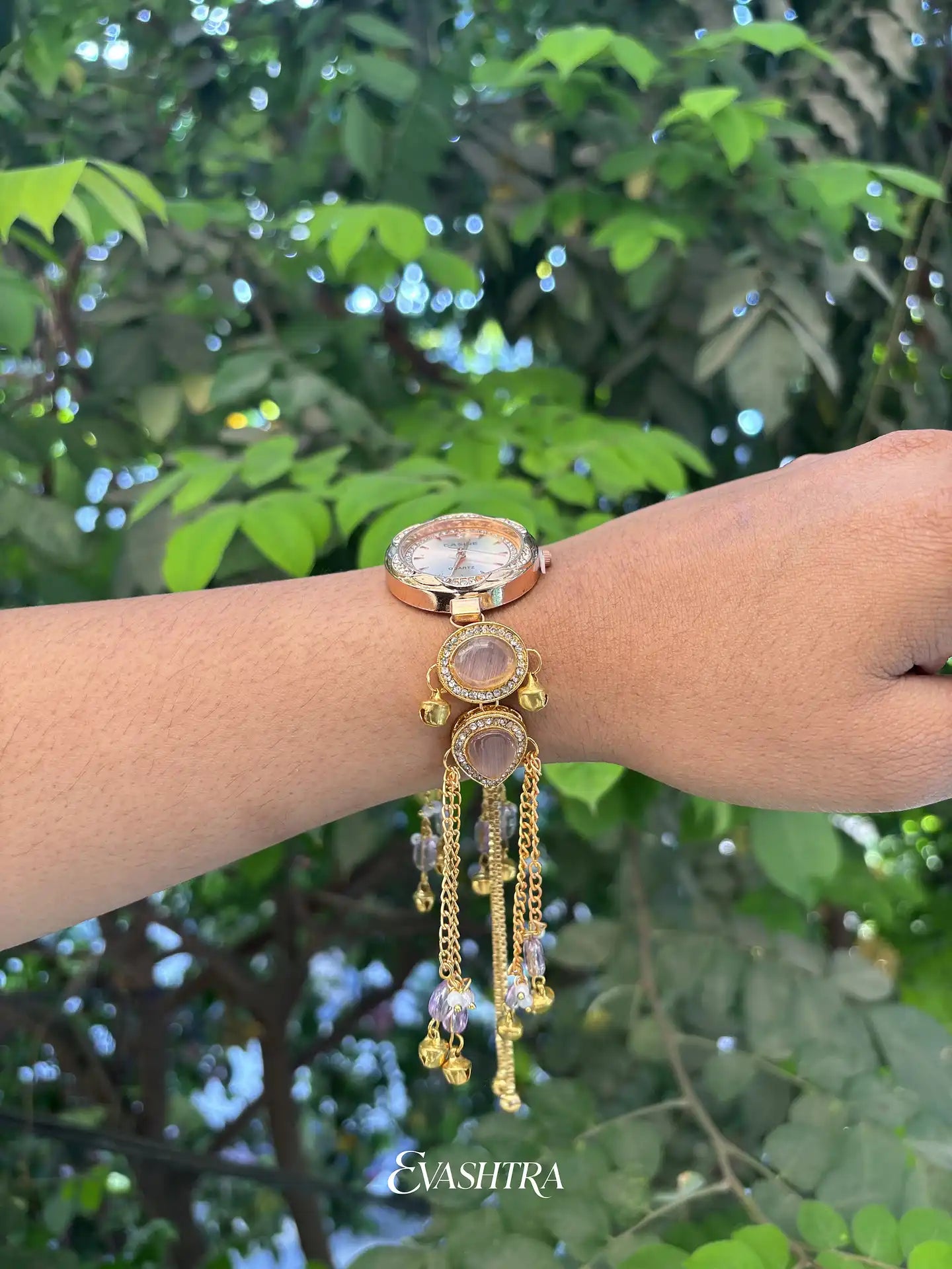 Gold wristwatch with dangling charms on a wrist against a green leafy background