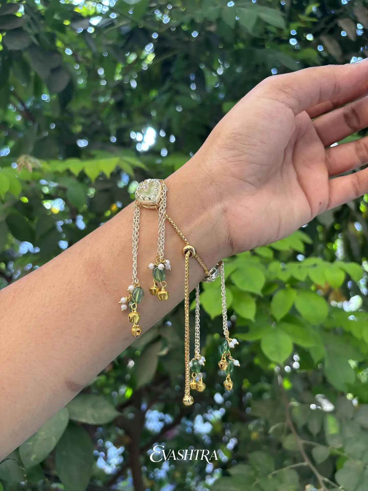 Hand wearing gold bracelets with green stones against a green leafy background
