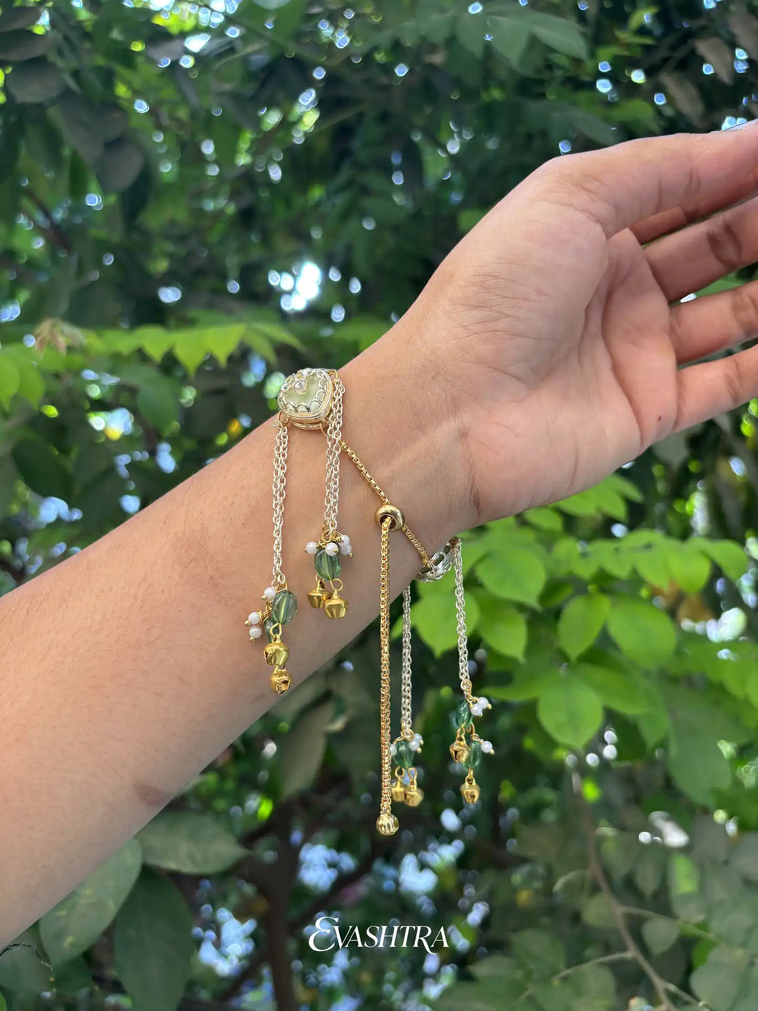 Hand wearing gold bracelets with green stones against a green leafy background