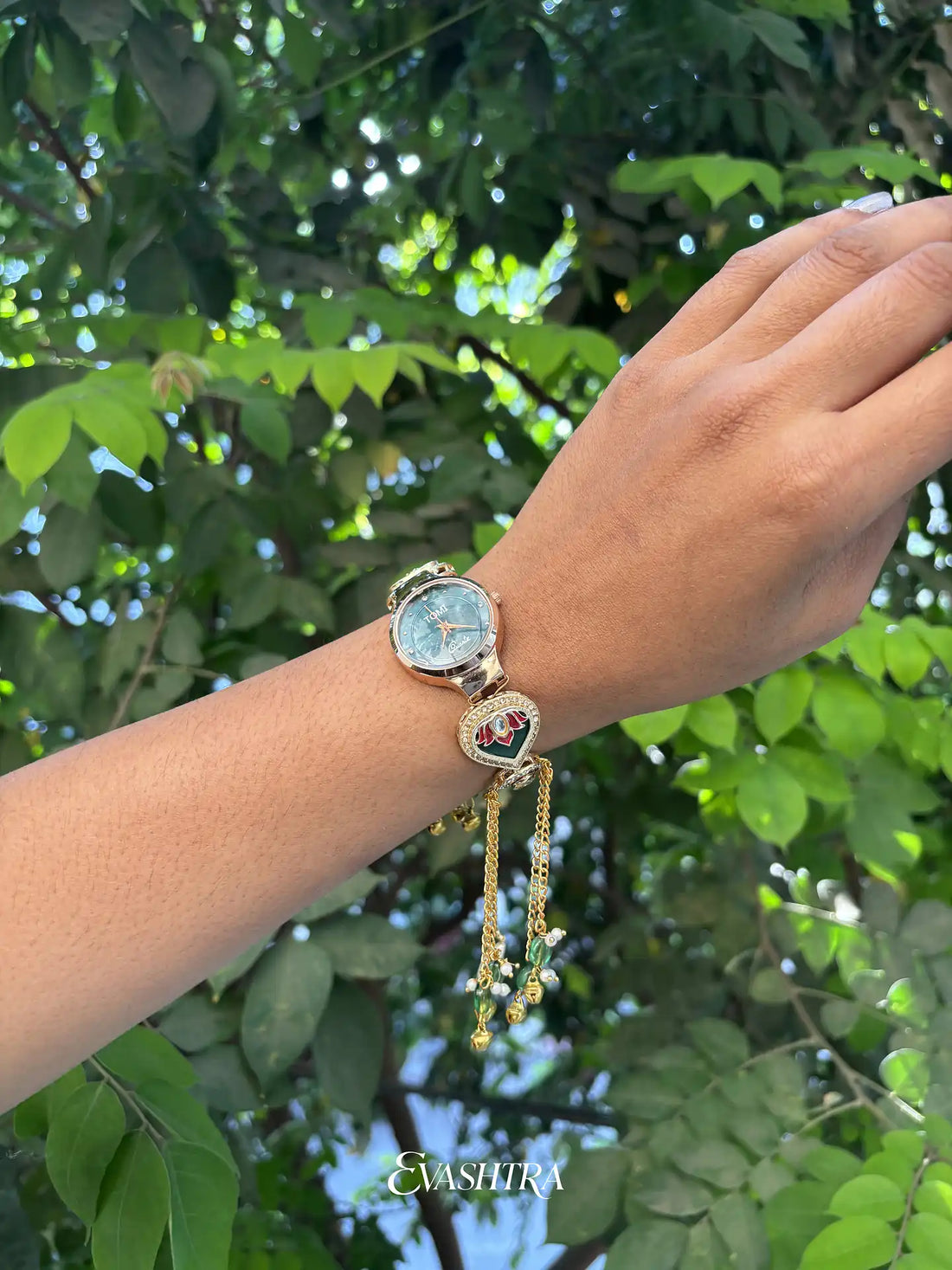 Hand wearing a gold bracelet with a watch face against a green leafy background
