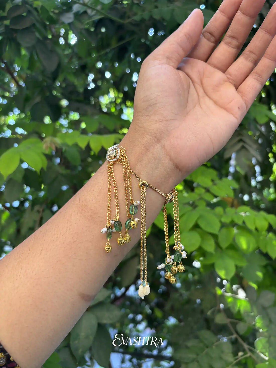 Hand wearing multiple gold bracelets with green leaves in the background