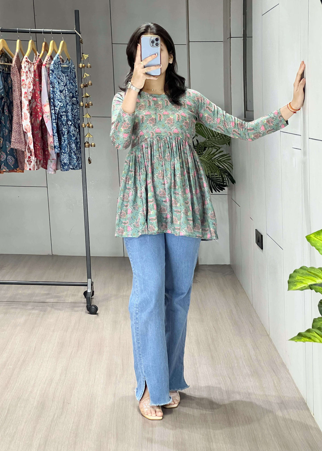 Person taking a mirror selfie wearing a floral top and blue jeans in a room with a clothing rack.