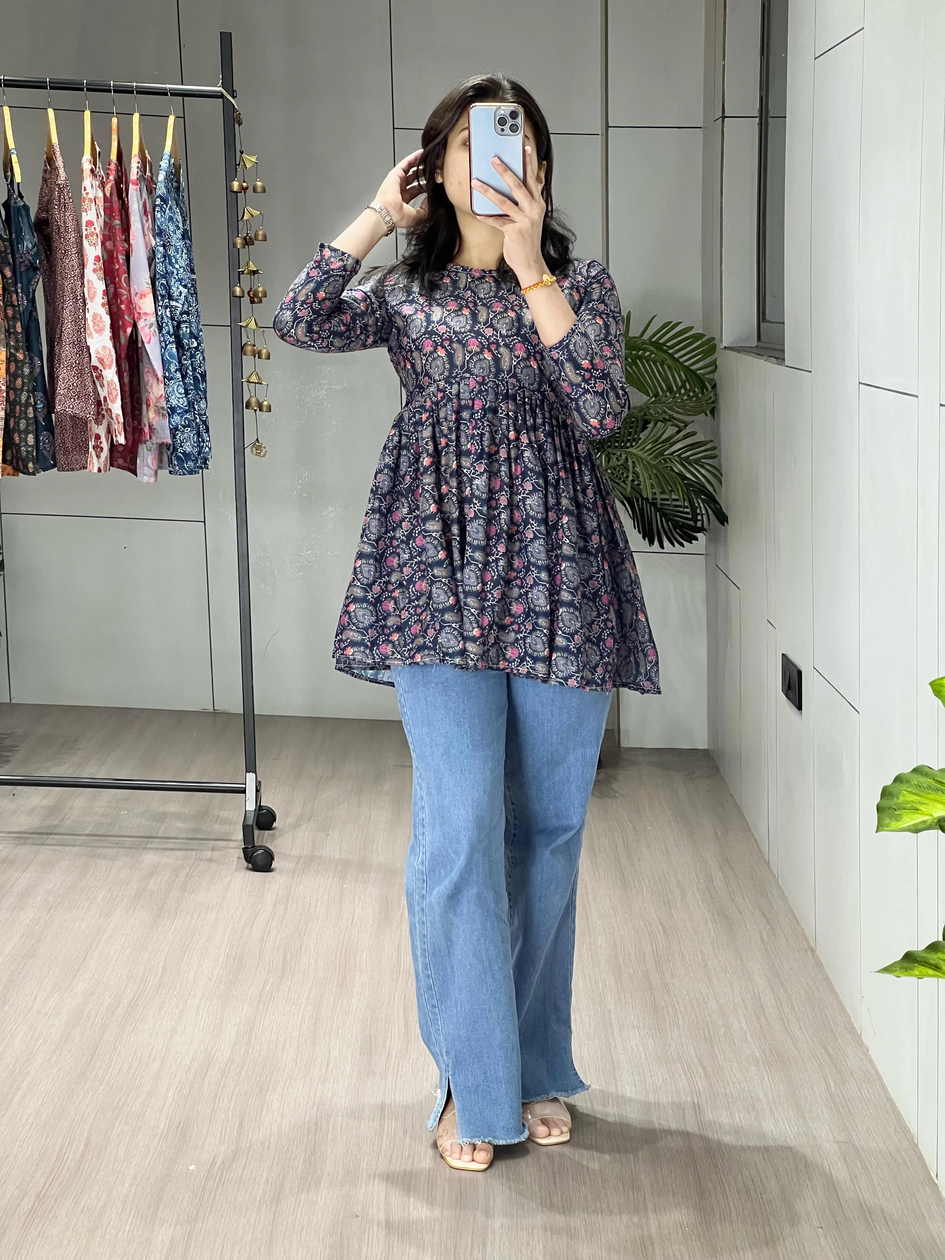 Person wearing a floral top and blue jeans taking a mirror selfie in a room with a clothing rack.