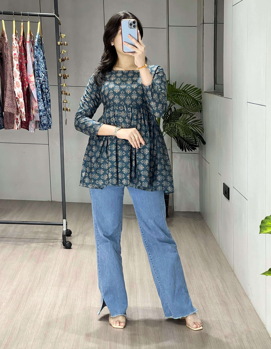 Woman taking a mirror selfie wearing a floral top and blue jeans in a room with a clothing rack.
