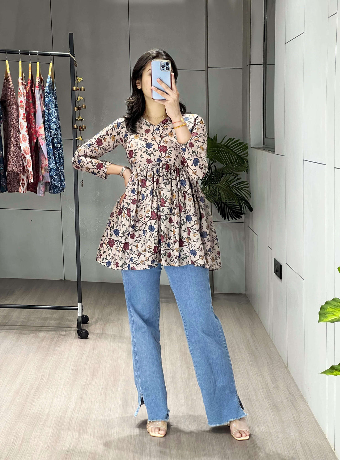 Person wearing a floral dress and blue jeans taking a mirror selfie in a room with a clothing rack.