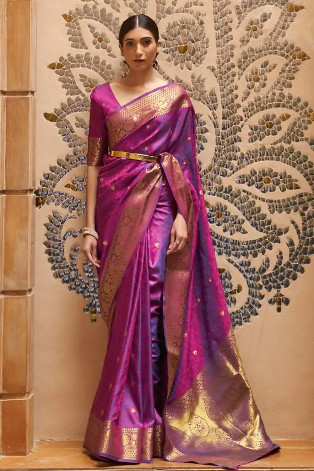 Woman wearing a vibrant purple saree with gold details against an ornate wall.