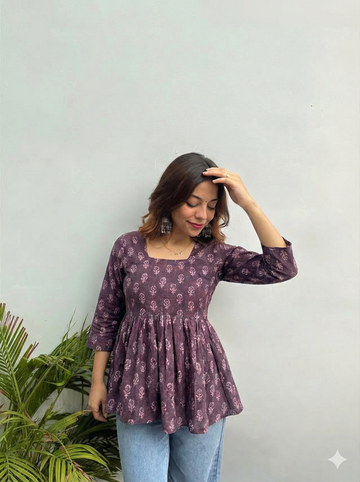 Woman wearing a purple floral blouse against a light green wall with a plant to the left.